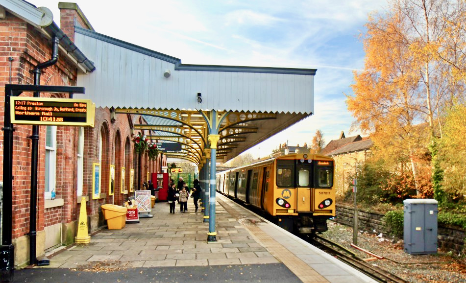 Ormskirk railway station with a Metro train - yellow front set on a blue sky with white clouds near the roof.